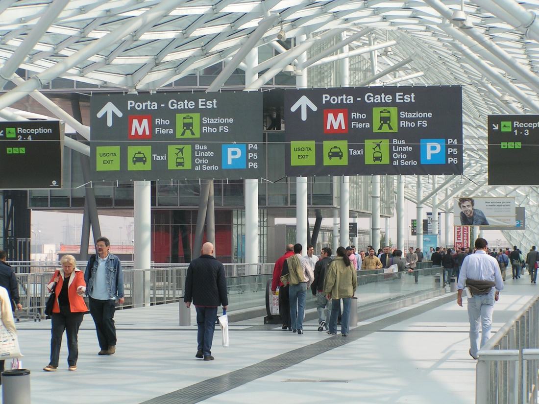 Interno di Fiera Milano a Rho: visitatori sotto la copertura di vetro e i cartelli per la metropolitana Rho Fiera
